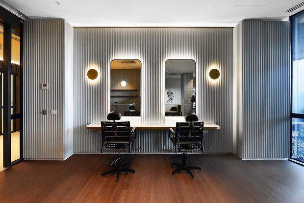 A refined contemporary salon or grooming workspace with two black styling chairs positioned at a long pale stone or solid-surface counter. The back wall is clad in vertically ribbed grey paneling and includes two rounded-rectangle mirrors, illuminated by paired circular wall sconces. Warm timber flooring, dark-framed glazing and doors, built-in shelving, and a glimpse of adjacent spaces complete the polished interior.