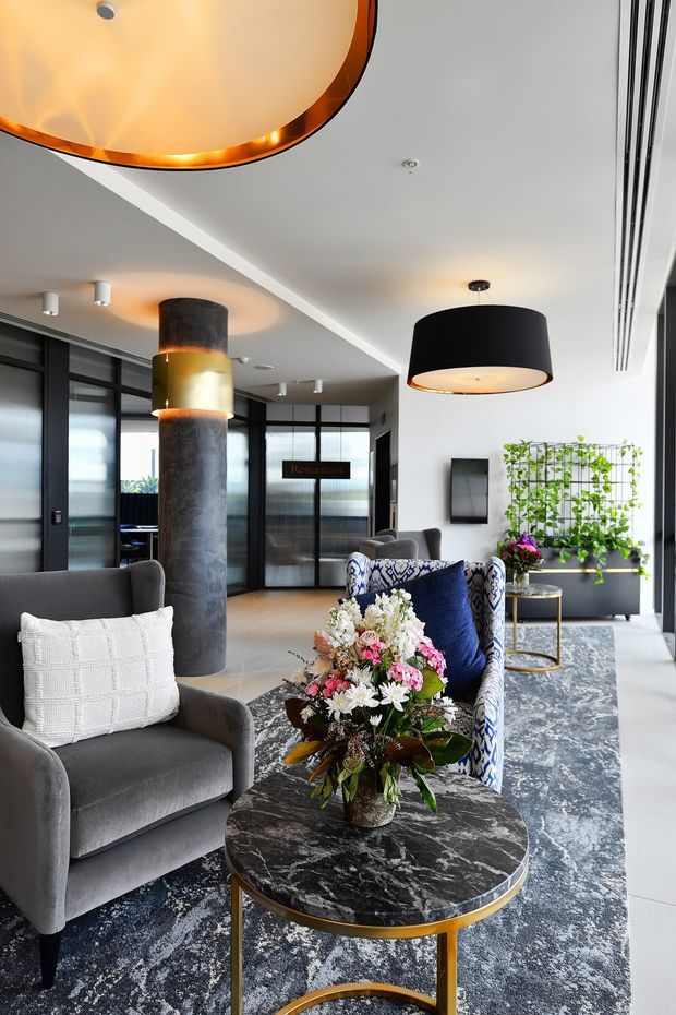 A refined contemporary lobby or reception lounge with grey upholstered armchairs, patterned blue accent seating, a round dark marble coffee table with brass edging, a large patterned rug, sculptural pendant lighting, black-framed glazed partitions, and a cylindrical textured feature column. A planted wall and reception area are visible in the background.