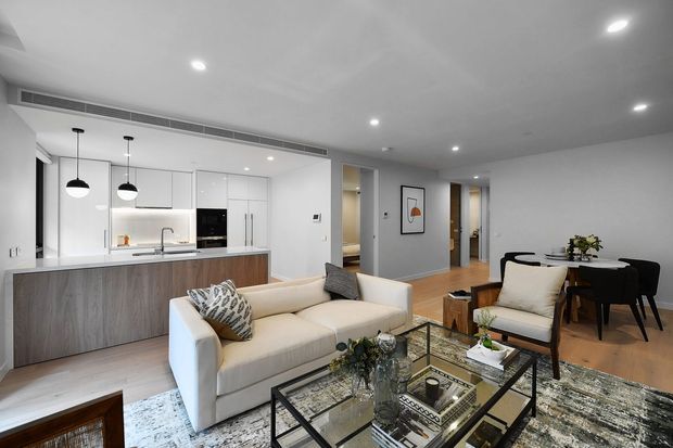 A bright, open-plan contemporary apartment interior combining a white kitchen with a timber-look island, a living area with a cream sofa and armchair, and a compact dining zone. Recessed ceiling lighting, pendant lights, pale timber flooring, neutral walls, and a large patterned rug create a clean, refined atmosphere. A hallway and additional rooms are visible beyond the main living space.