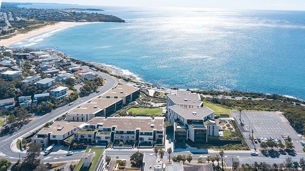 Aerial view of a coastal urban precinct, with modern multi-storey buildings, landscaped courtyards, roads and a large parking area beside a sandy beach and expansive blue ocean.