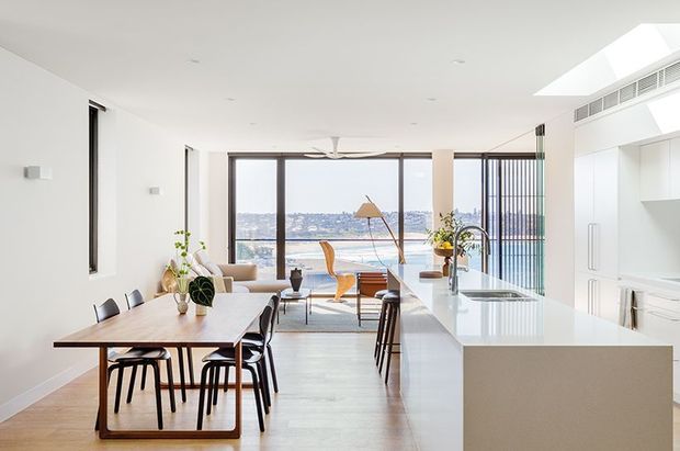 Bright open-plan kitchen and dining area with a long pale timber dining table, black dining chairs, a large white kitchen island with integrated sink, extensive floor-to-ceiling glazing, and a waterfront view beyond. A skylight and linear ceiling ventilation add to the airy contemporary interior.