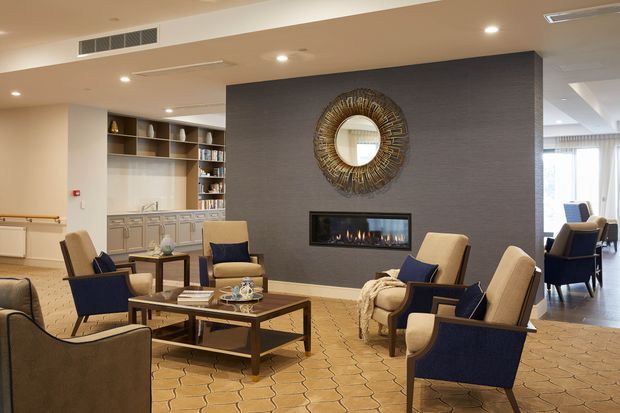 A refined communal lounge with multiple upholstered armchairs arranged around low wooden coffee tables. A dark textured feature wall contains a linear fireplace and large decorative round mirror, while built-in shelving and cabinetry form a library or refreshment area in the background. The space is warmly lit and opens toward a bright dining or sitting area.