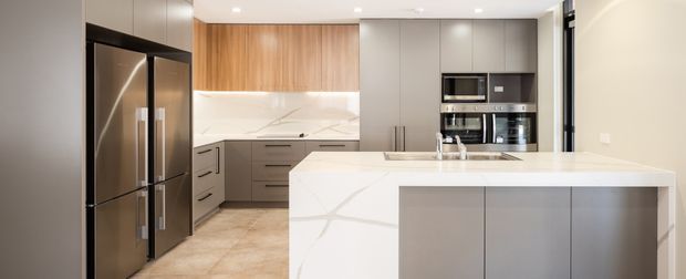 Contemporary kitchen featuring a large white stone island with waterfall edges, grey flat-panel cabinetry, warm timber overhead cabinetry, integrated stainless-steel appliances, and a pale tiled floor. The bright, open layout has recessed ceiling lights and a clean, refined finish.