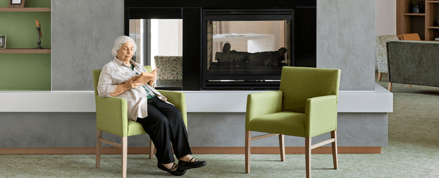 A contemporary living room centered on a built-in black fireplace set into a grey textured wall, with a white horizontal ledge and green built-in shelving. Two lime-green upholstered armchairs with timber legs sit on a pale green rug, while an older person reads in one chair. A partial adjoining seating area is visible at right.