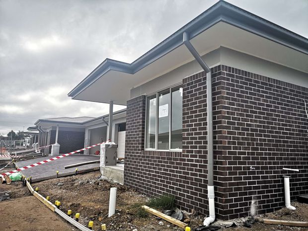 Exterior view of a newly constructed single-storey residential building, showing dark brick walls, white-framed windows, projecting eaves, a low-pitched gable roof, and exposed drainage pipes. The surrounding ground is unfinished with construction materials and safety tape visible.