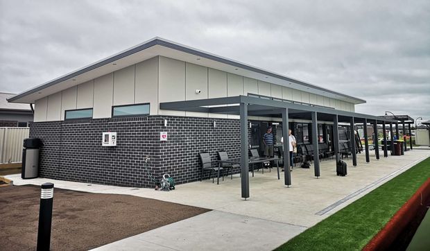 Exterior view of a contemporary single-storey building with a low-pitched roof, pale vertical wall cladding, and a dark brick masonry base. A covered outdoor walkway or verandah with slender dark metal posts extends along the facade, with outdoor seating and several people beneath it. The foreground includes concrete paving, landscaped garden beds, bollard lighting, and artificial turf. Overcast daylight.