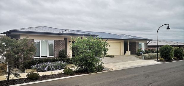 Single-storey contemporary suburban residential building viewed from the street, with a low-pitched metal roof, rendered and brick exterior walls, broad covered entry and garage, concrete driveway, landscaped front gardens, and a distinctive curved black exterior light fitting. Overcast conditions create a muted, neutral presentation.