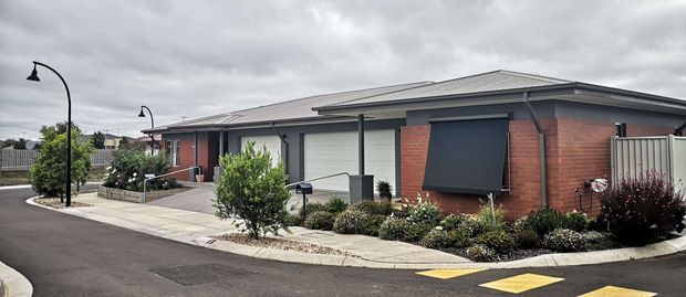 Exterior view of a modern single-storey residential development with low-pitched hipped roofs, red brick walls, dark grey metal-framed openings and garage doors, and landscaped front paths along a paved street. The overcast sky and street furniture create a subdued contemporary suburban setting.