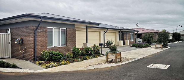 A street-level view of a low-rise modern residential development with several attached single-storey dwellings. The homes feature dark low-pitched rooflines, brick and light rendered exterior walls, white-framed windows, concrete pedestrian paths, small landscaped garden beds, and a paved internal roadway under an overcast sky.