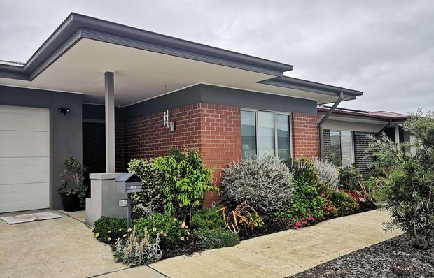 Single-storey contemporary residential facade viewed from the front, featuring a broad low-pitched roof with deep eaves, dark grey painted wall sections, red brick feature walls, aluminium-framed windows, an attached garage, and a landscaped front garden with a concrete walkway.