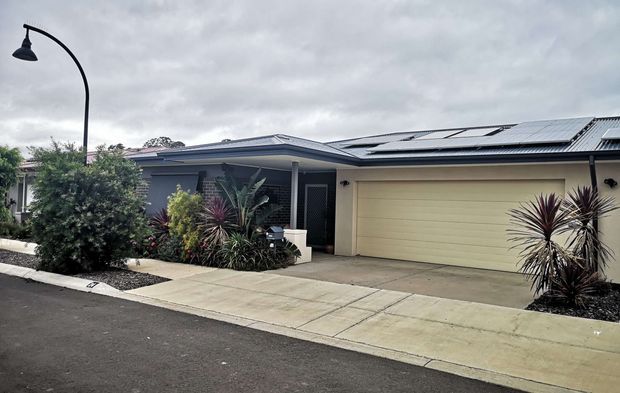 Single-storey contemporary suburban home viewed from the street, with a low-pitched roof fitted with solar panels, a broad integrated double garage, covered entry, dark-framed glazing, and drought-tolerant landscaped planting along the driveway and frontage. The overcast sky and dark pavement create a subdued exterior palette.
