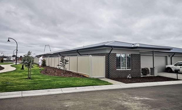 Single-storey contemporary suburban dwelling viewed from the street, with a low-pitched hipped metal roof, dark brick feature walls, light rendered walls, and a covered carport or garage at right. The property has a newly landscaped front lawn, young trees, garden beds, a pale privacy fence, concrete footpaths, and a broad paved street under an overcast sky.