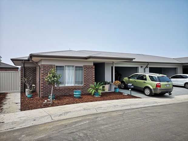 Single-storey suburban brick house viewed from the street, with a low-pitched metal roof, covered entry porch, front garden bed, driveway and attached double garage. The facade combines dark red-brown brickwork with light rendered or painted sections and white-framed windows.