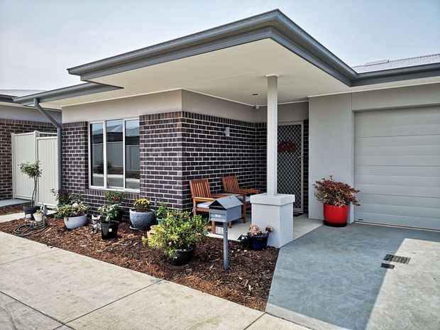 Exterior view of a contemporary single-storey townhouse or residential unit with a flat, projecting porch roof, dark brickwork, white rendered walls, a covered entry patio, garage, paved driveway and landscaped garden beds with potted plants. Two timber outdoor chairs and a small table create a compact front seating area.