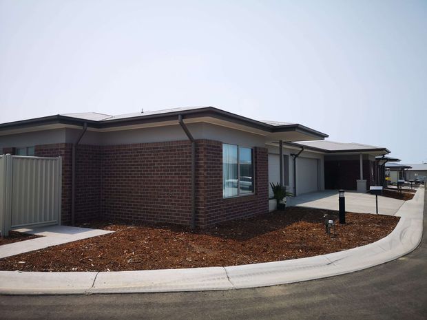 Single-storey contemporary brick house viewed from the street, with a low-pitched roof, attached covered carport, concrete driveway and sparse landscaped frontage. Dark brick walls contrast with pale rendered or panelled sections and light metal garage/carport elements.