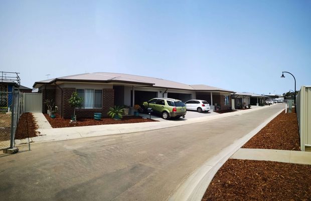 A single-storey contemporary residential development viewed from a broad internal driveway. The row of attached or closely spaced homes has low-pitched metal roofs, brick and rendered facades, covered porch or carport areas, front windows, small landscaped garden beds, and several parked cars. Concrete paving and dark mulch dominate the foreground, with a clear blue sky and simple black street lighting.