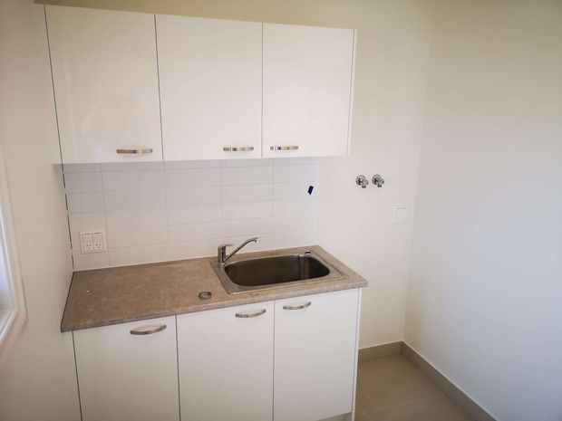 Compact, unfurnished kitchenette or utility kitchen with white overhead and base cabinetry, a stainless-steel sink and mixer tap, a mottled brown benchtop, and a white rectangular tiled splashback. The room has plain white walls, beige floor tiles, and minimal detailing.