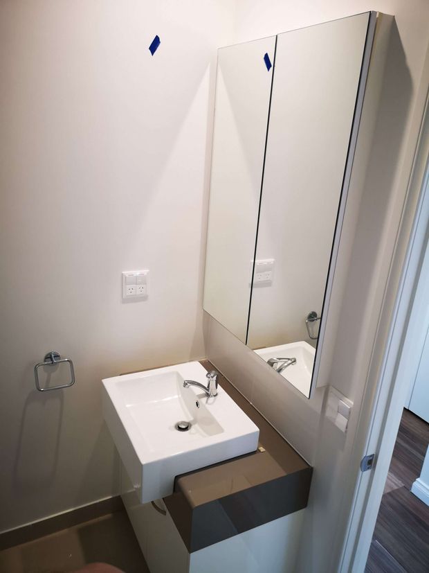Compact contemporary bathroom vanity area with a white rectangular basin, chrome tapware, mirrored wall cabinet, neutral painted walls, and dark built-in vanity cabinetry. A doorway opens to a timber-finished adjacent space.