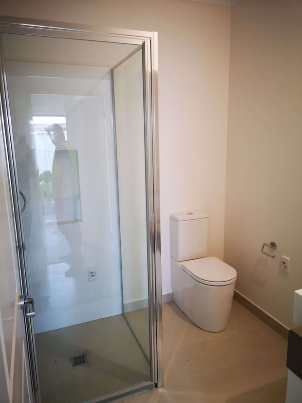 A clean, contemporary bathroom with a large framed glass shower enclosure on the left and a white toilet beside it. The room has pale beige painted walls, light neutral floor tiles, glossy white shower wall tiles, and chrome shower and bathroom hardware. A partial vanity is visible at the right edge. The shower glass reflects the photographer and doorway.