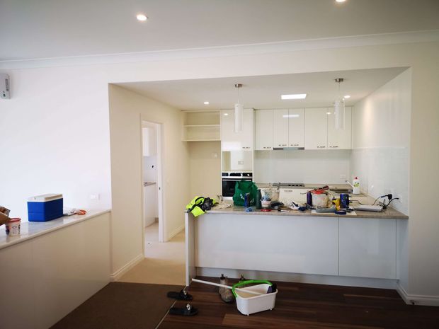 A bright, recently fitted open-plan kitchen viewed from an adjoining living or dining area. The kitchen has white upper cabinetry, pale walls, a light stone-look benchtop, a pale blue-grey island with integrated cabinetry, recessed ceiling lights, and dark timber flooring. Cleaning supplies and renovation-related clutter are visible on the counters and floor.