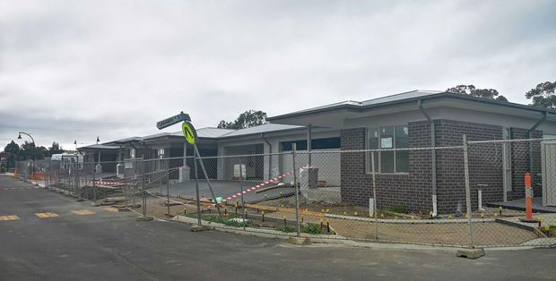Street-level exterior view of a low-rise contemporary building under construction, with dark brick walls, a shallow metal roof, broad windows, and a fenced construction zone. The surrounding paved roadway, temporary safety barriers, signage, and construction materials are visible.