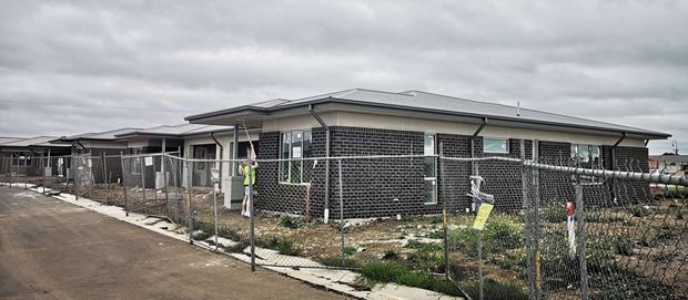 Single-storey contemporary low-rise building under construction, with dark brick masonry walls, a broad shallow hipped roof, and multiple small horizontal windows. The site is enclosed by temporary chain-link fencing, with unfinished landscaping and paved access paths visible under an overcast sky.