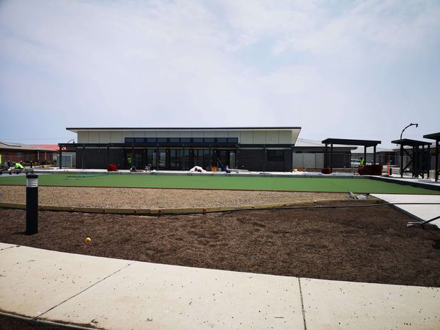 Wide exterior view of a low-rise contemporary recreational or commercial facility, with a broad flat roof, dark glazed facade, covered outdoor structures, paved paths, and landscaped or unfinished ground in the foreground. The setting appears to include a large green synthetic playing surface and outdoor activity areas.
