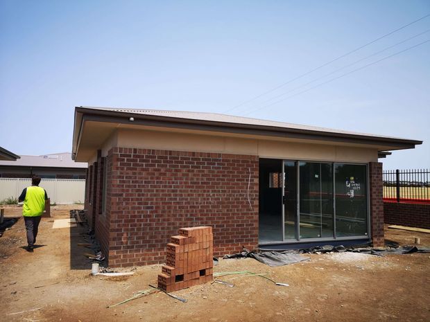 Single-storey brick building under construction, with red brick walls, a low-pitched metal roof with broad eaves, and large dark-framed glazed sliding doors at the front. Exposed dirt surrounds the structure, with stacked bricks, construction materials and unfinished landscaping visible. The scene is photographed from outside in bright daylight.