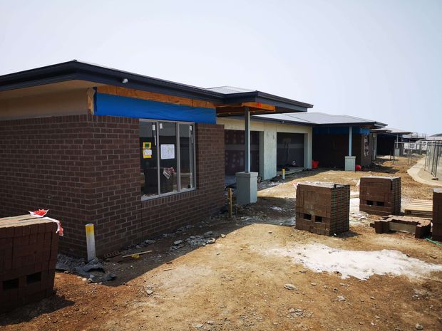 Exterior construction-stage view of a low-rise modern residential or institutional building with dark brick walls, flat or low-pitched rooflines, exposed timber framing, windows and doors, and unfinished dirt and paving around the structure. Construction materials and pallets are visible in the foreground.