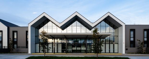 Front exterior view of a contemporary commercial or institutional building with a distinctive repeated zigzag roofline. The facade combines dark vertical cladding, grey masonry at the base, white angular roof frames, extensive floor-to-ceiling glazing, and a deep covered entrance canopy. Young trees and a paved forecourt sit in front of the building under a clear sky.