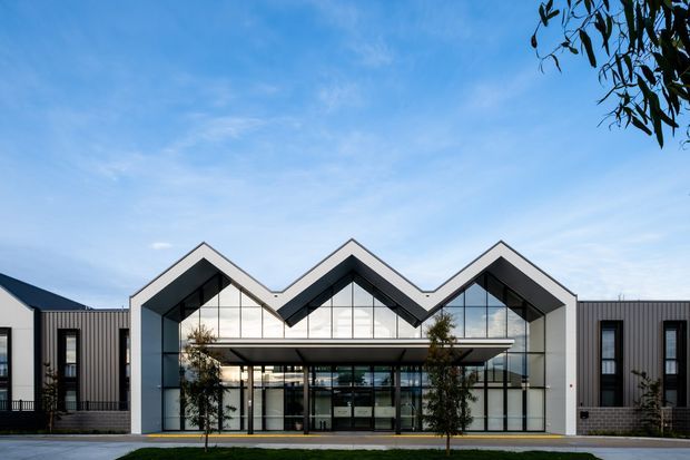 Front exterior view of a contemporary multi-unit building with a striking repeated zigzag gable roofline, dark vertical metal cladding, and extensive floor-to-ceiling glazing. A broad covered entrance canopy projects over the central glazed lobby, with landscaped foreground and a clear sky beyond.