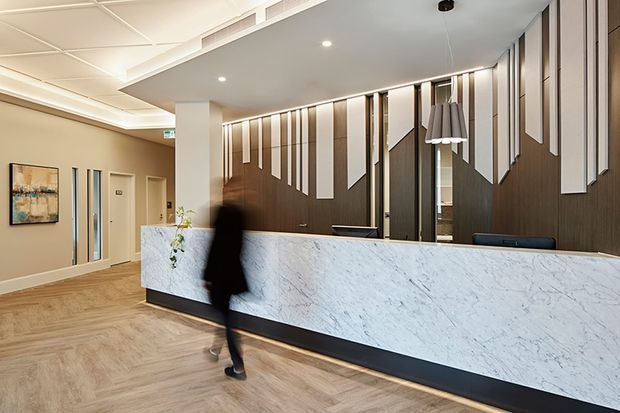A refined commercial reception lobby with a long white veined stone reception desk, dark timber feature wall, and vertically arranged geometric light panels behind the desk. The space has warm herringbone timber flooring, beige walls, recessed ceiling lighting, a suspended pendant, and a corridor leading away at the left. A blurred person is walking in front of the desk.