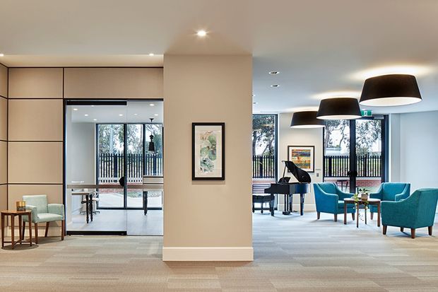 A bright, refined open-plan interior resembling a hotel or commercial lounge. The foreground features a neutral carpeted floor and a large beige partition wall with integrated geometric panel detailing and a framed artwork. Beyond it is a glazed transition to a lounge area furnished with teal armchairs, small tables, a grand piano, and oversized black pendant lights. Large windows provide views of trees and a black balustrade outside.