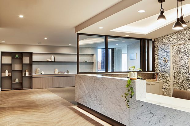 A refined contemporary reception or hospitality space featuring a large marble-clad front counter, dark timber cabinetry and open shelving, black-framed internal glazing, recessed ceiling lighting, pendant lights, and a striking patterned feature wall. The warm neutral palette and layered lighting create an upscale, welcoming atmosphere.