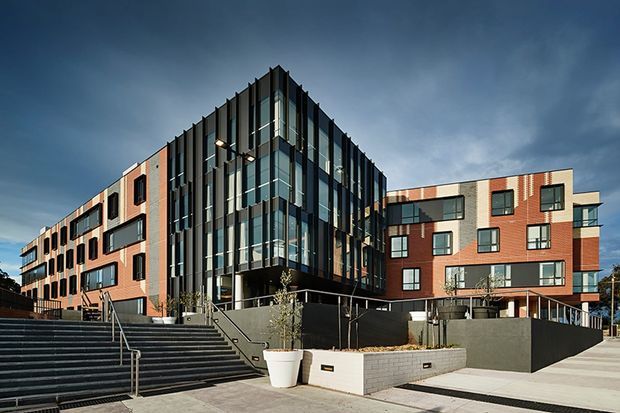 Exterior architectural photograph of a contemporary multi-storey apartment or mixed-use building complex. The composition features a prominent glazed central corner volume with dark vertical metal fins, flanked by warm red-brick wings with large black-framed windows and balconies. Broad concrete steps, paved walkways, retaining walls, and landscaped planters define the foreground. The building has a crisp modern form under a dramatic cloudy sky.