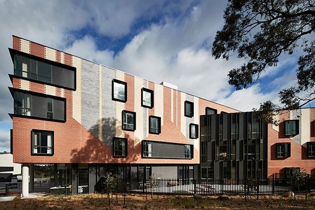 Exterior view of a contemporary multi-storey brick building with a strong patterned facade, black-framed windows, projecting glazed bays, and a dark vertical glazed entrance or circulation volume. The building is set behind a black metal fence, with a partly cloudy sky and mature tree nearby.