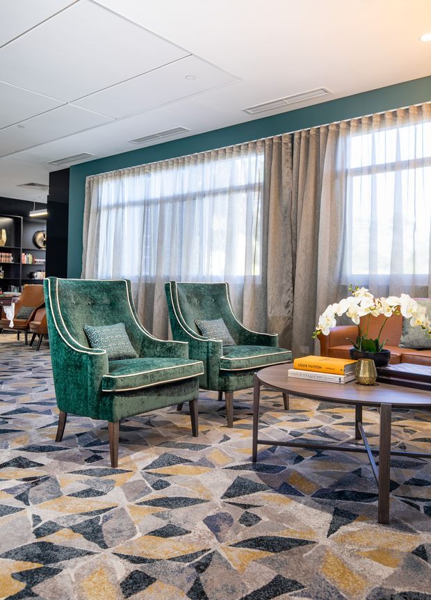 A stylish hotel or commercial lounge seating area with two emerald-green velvet wingback armchairs, patterned carpet, a round wooden coffee table, sheer and textured drapery, and a teal feature wall. Built-in shelving and additional seating are visible in the background, creating a refined, eclectic atmosphere.