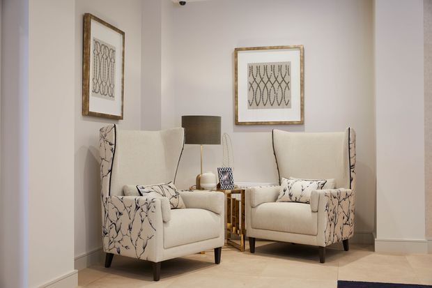 A refined sitting area with two matching upholstered wingback armchairs in cream fabric with botanical-patterned side panels, arranged around a small side table and floor lamp. Framed monochrome artwork hangs on a warm light wall, with pale tiled flooring and restrained decorative styling.