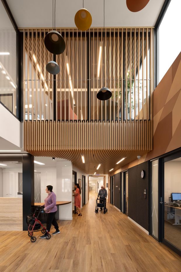 A bright, contemporary multi-level commercial interior viewed from a wide circulation corridor. The space features warm timber-look flooring, a dramatic timber slatted mezzanine and ceiling soffit, full-height glazing, dark-framed office fronts, linear pendant lighting, and a sculptural faceted timber-toned wall. Several people are using the accessible hallway, which opens toward reception and glazed workspaces.