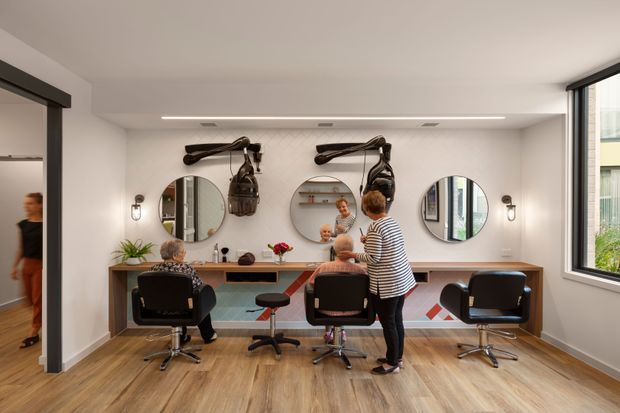 A bright, contemporary hair salon interior with three styling stations arranged along a long timber-toned counter. Each station has a round wall mirror, salon chair, and overhead sculptural hairdryer fixtures, with additional decorative lighting and plants. The room features pale walls, warm wood-look flooring, large windows, and a clean, refined commercial fit-out.