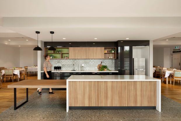 A contemporary open-plan commercial kitchen and dining space. The foreground features a large island with light timber cabinetry, a pale stone benchtop, and a dark recessed base. Behind it is a linear kitchen with timber overhead cabinetry, black lower cabinetry, patterned tile splashback, open green shelving, integrated appliances, and black pendant lights. Timber dining tables and chairs are visible on both sides, while polished concrete or terrazzo flooring spans the room.