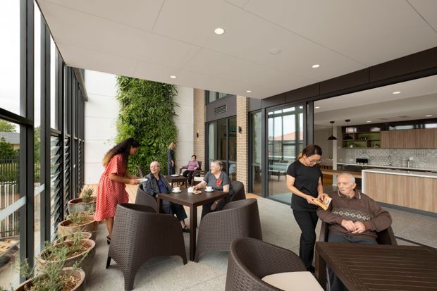A bright, contemporary communal dining and lounge area with full-height glazing, outdoor planters, a planted vertical wall, and several people gathered around café-style tables. A modern kitchen and service counter are visible through the open glazed partition on the right. The space has a light tiled floor, dark woven seating, timber-look cabinetry, and a white recessed ceiling with downlights.