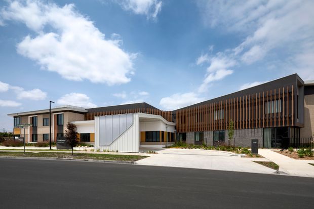 A contemporary multi-wing building viewed from across a newly paved street. The facade combines dark horizontal roof forms, extensive vertical timber screening, pale masonry or concrete elements, and large glazed sections. A prominent white angular entrance volume projects from the centre, with landscaped planting and broad concrete driveways in front. Blue sky and clouds form the background.
