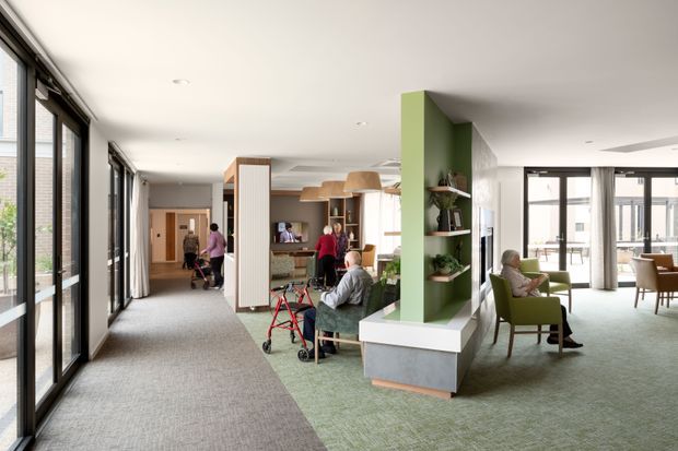A bright, open-plan communal living and lounge area with extensive floor-to-ceiling glazing along the left side, green carpet, a large green built-in shelving and partition wall, and several seating zones. Older adults are using the space, with a visible walking aid and communal dining or activity areas in the background. The interior has a calm, contemporary residential-care or community setting with abundant natural light and an airy layout.
