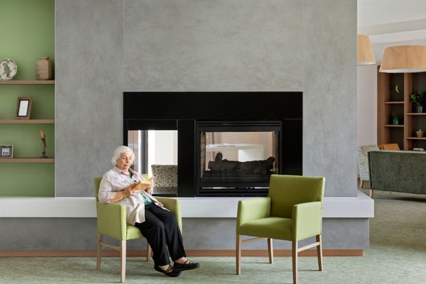 A refined living room centered on a large black fireplace set into a tall grey textured feature wall. Two lime-green upholstered armchairs sit on a pale rug, with an elderly person seated in one. Built-in open shelving painted green appears on the left, while timber-toned shelving and a second seating area are visible through the opening on the right. The room has a calm, contemporary feel with strong contrasting green, grey, black, and timber elements.
