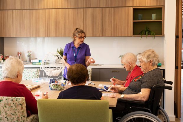 A bright communal activity and dining area in an aged-care or assisted-living setting. Several elderly people are seated around a table with a staff member, while a large wall of warm timber cabinetry, white backsplash, dark lower cabinetry, and an open display shelf forms the backdrop. The space includes accessible seating and a wheelchair, with a calm, welcoming institutional-interior character.