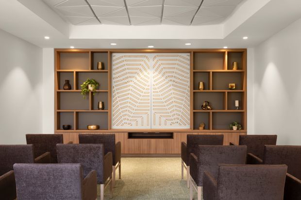 A refined contemporary lounge or reception area with rows of upholstered armchairs facing a built-in timber media or feature wall. Symmetrical open shelving displays small decorative objects and plants, while the central pale geometric wall panel provides a focal point above a low timber cabinet. Recessed lighting and a patterned acoustic ceiling create a polished, softly lit interior.