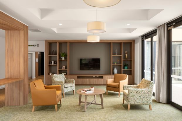 Bright, refined living room with a warm timber built-in media wall and shelving, recessed ceiling details, pendant lighting, and large floor-to-ceiling glazed doors with curtains. The room is arranged with caramel armchairs, patterned lounge chairs, a round timber coffee table, and a muted textured rug, creating an inviting contemporary interior.