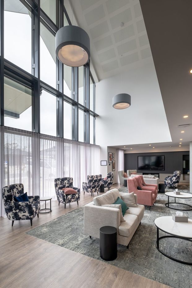 A spacious, high-ceilinged contemporary lounge with a dramatic double-height glazed facade, sheer curtains, patterned armchairs, blush-pink lounge chairs, neutral sofas, round coffee tables, and a large textured rug. The room features dark metal window framing, a dark vertical-panel media wall, oversized cylindrical pendant lights, recessed ceiling lighting, and pale timber flooring.
