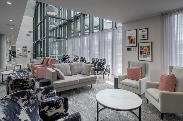 A bright, contemporary lounge or waiting area with multiple upholstered armchairs and sofas arranged around low round coffee tables. The space features a largely neutral palette with blush-pink seating accents, patterned navy and cream chairs, full-height sheer curtains, polished dark-framed floor-to-ceiling glazing, recessed ceiling lighting, pendant lights, framed abstract artwork, and a large pale rug. The glazing creates a tall, airy atrium-like feel, with a corridor or adjacent interior visible beyond.